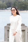 Load image into Gallery viewer, A woman wearing a matching cream-colored two-piece lounge set consisting of a long-sleeve top and wide-leg pants, standing outdoors by a waterfront with a blurred background of water, trees, and buildings.
