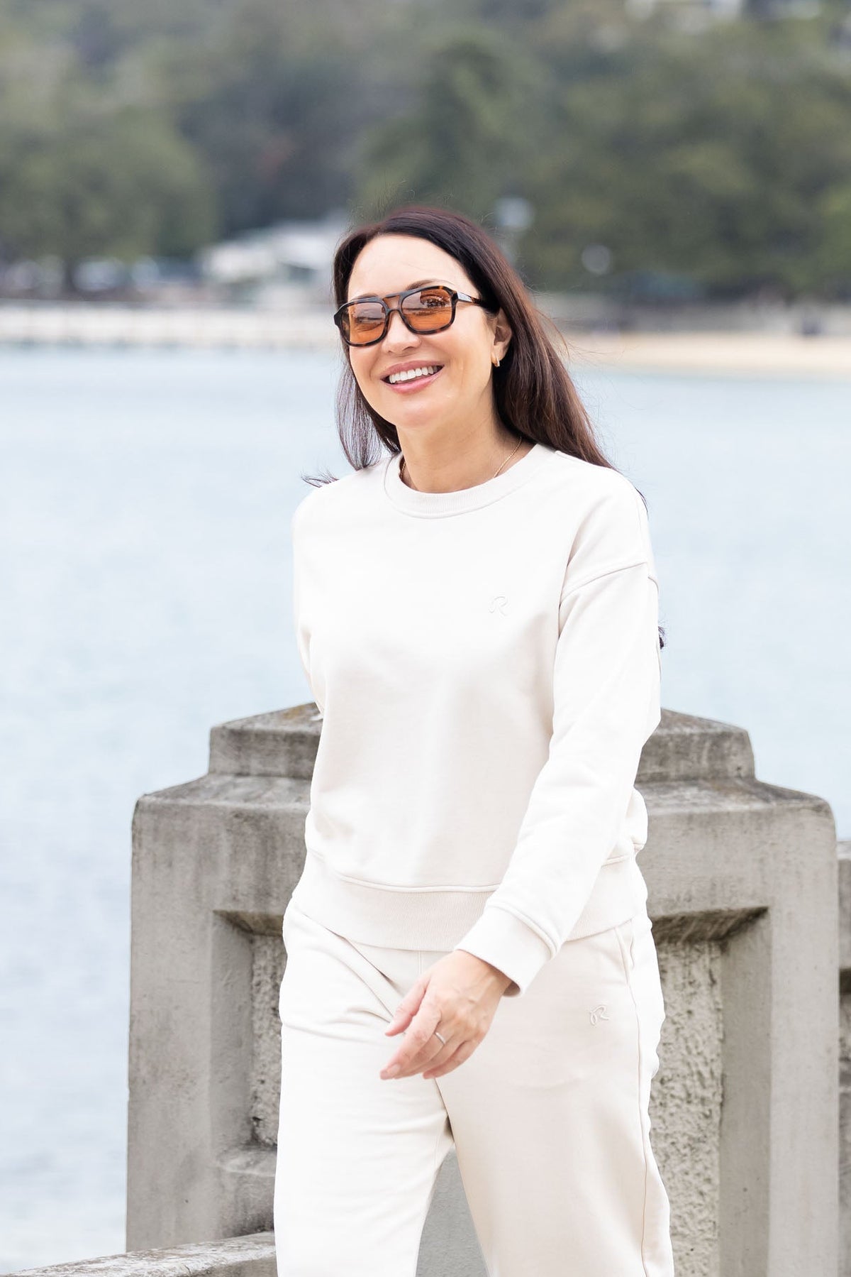 A woman wearing a matching cream-colored two-piece lounge set consisting of a long-sleeve top and wide-leg pants, standing outdoors by a waterfront with a blurred background of water, trees, and buildings.