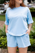 Load image into Gallery viewer, A woman wearing a light blue short-sleeve t-shirt and matching shorts set, styled with black sunglasses, standing outdoors in front of green plants.
