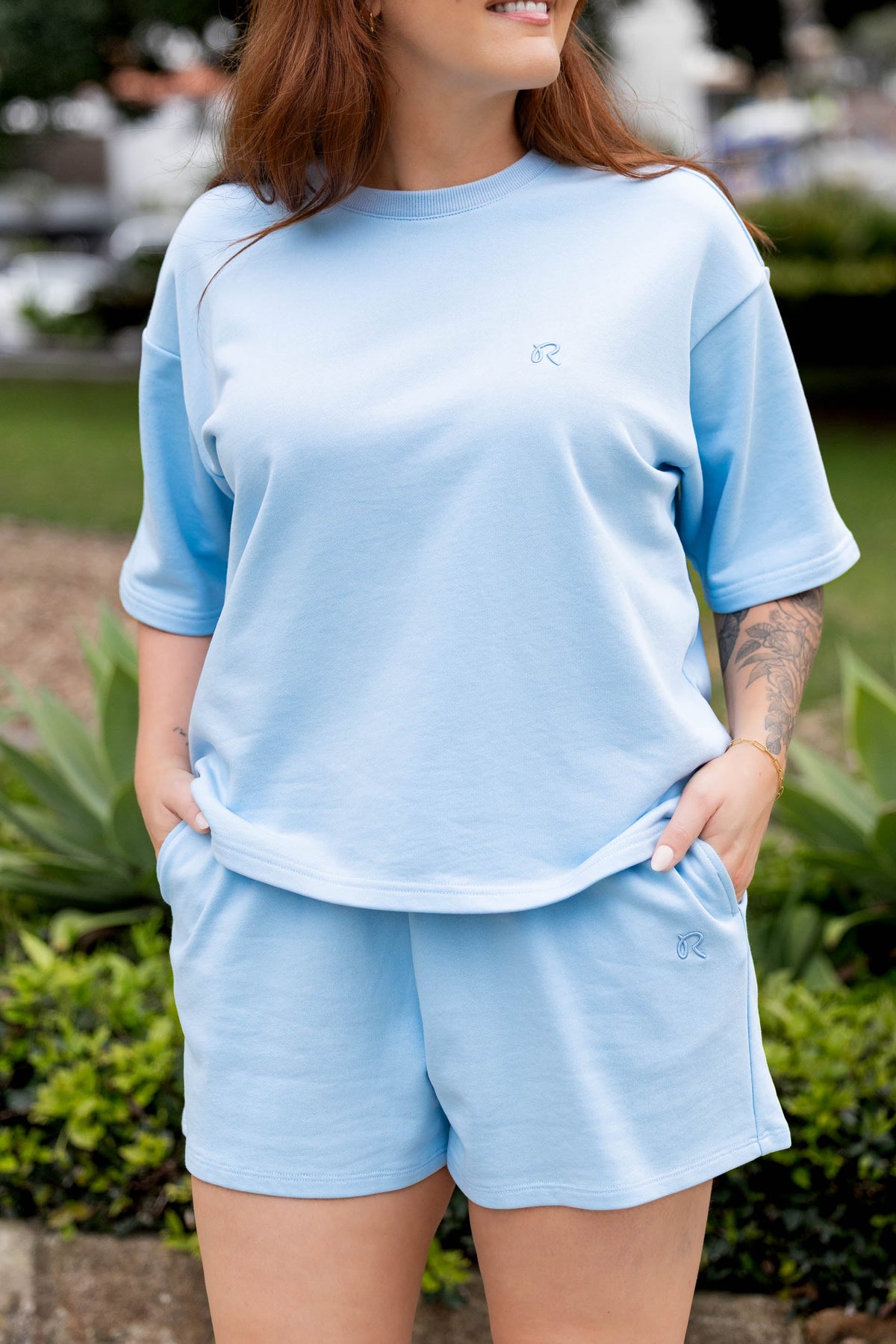 A woman wearing a light blue short-sleeve t-shirt and matching shorts set, styled with black sunglasses, standing outdoors in front of green plants.